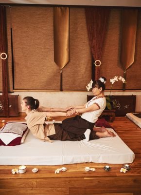 Woman Thai massage professional sitting with knees on female butt and pulling her by hands on mattress