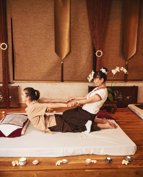 Woman Thai massage professional sitting with knees on female butt and pulling her by hands on mattress